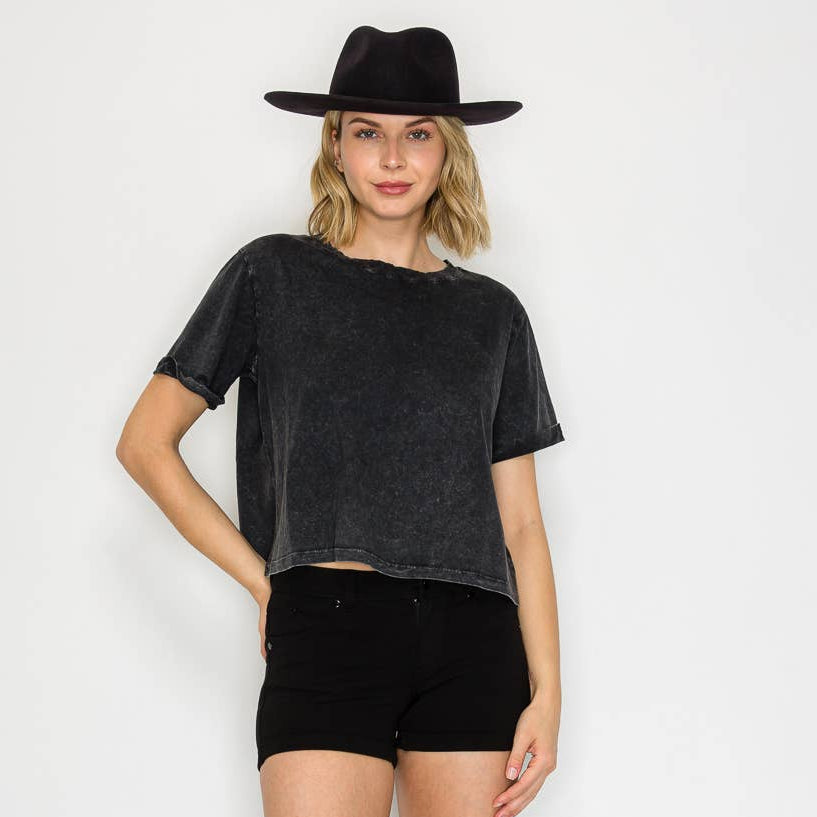 A female model wearing a black mineral-washed oversized t-shirt with a crew neckline, cut-off at the abdomen, paired with black shorts and black boots, accessorized with a black hat.